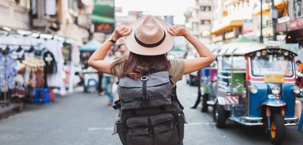 A woman with a large backpack and hat stands in a lively street market, facing away. Colorful tuk-tuks are in the background, conveying a sense of adventure.