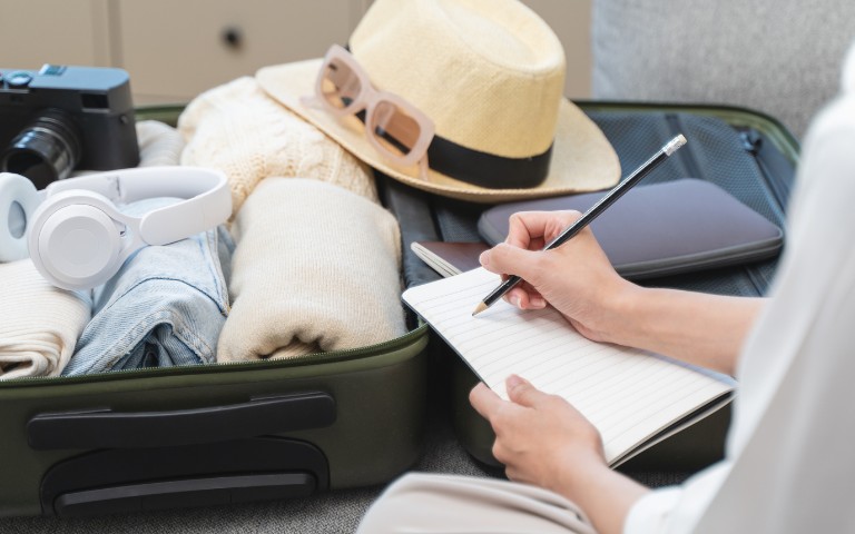 Person writing in a notebook next to an open suitcase filled with clothes, headphones, a camera, hat, and sunglasses, conveying a travel preparation vibe.