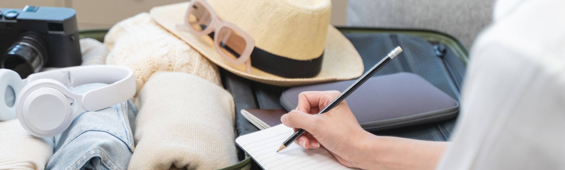 Person writing in a notebook next to an open suitcase filled with clothes, headphones, a camera, hat, and sunglasses, conveying a travel preparation vibe.