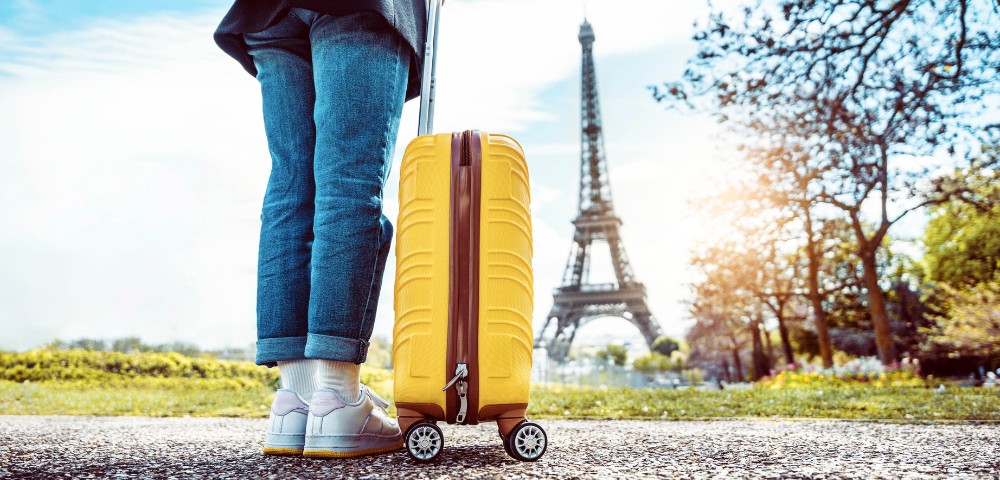 Traveler with a yellow suitcase stands on a path near the Eiffel Tower, wearing casual jeans and sneakers. The scene conveys a sense of adventure and excitement.