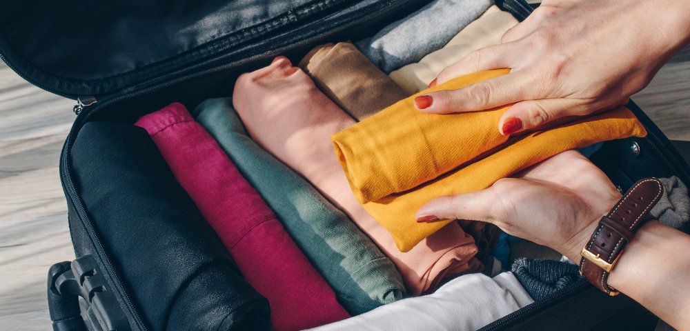 Hands are neatly packing colorful, rolled clothes into an open suitcase on a light wood floor. The scene conveys organization and travel readiness.