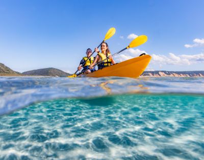 Two people kayaking in a bright yellow kayak on clear turquoise water, with yellow paddles. The sky is blue and the distant shore is visible.