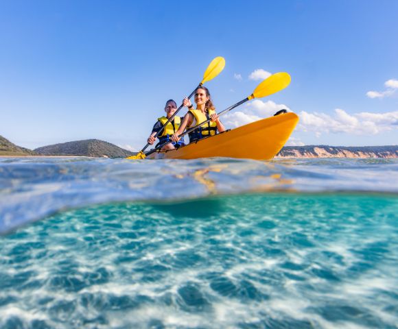 Two people kayaking in a bright yellow kayak on clear turquoise water, with yellow paddles. The sky is blue and the distant shore is visible.
