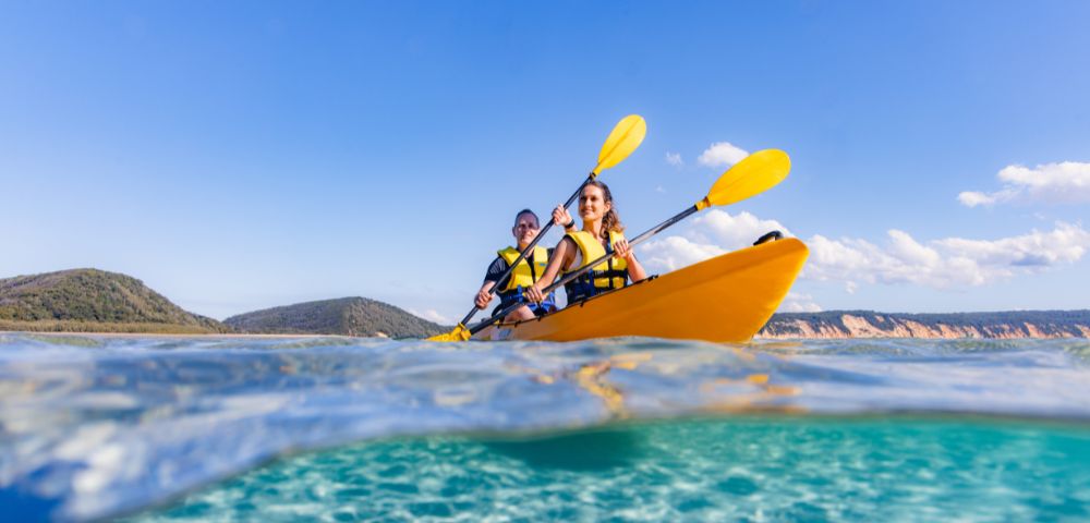Two people kayaking in a bright yellow kayak on clear turquoise water, with yellow paddles. The sky is blue and the distant shore is visible.