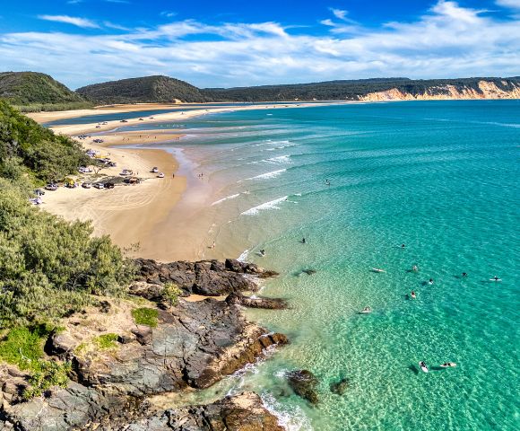 Aerial view of a scenic beach with turquoise waves gently lapping the shore. Swimmers dot the water, while lush greenery borders the sandy coast.