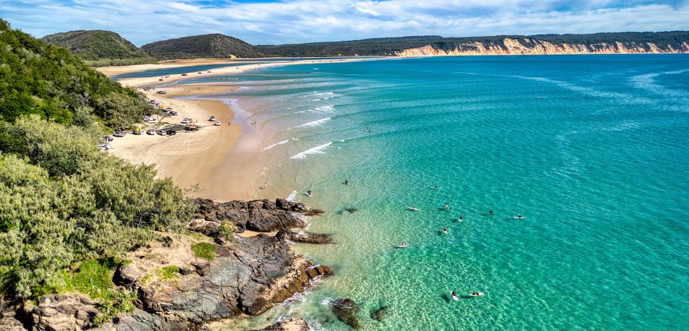 Aerial view of a scenic beach with turquoise waves gently lapping the shore. Swimmers dot the water, while lush greenery borders the sandy coast.