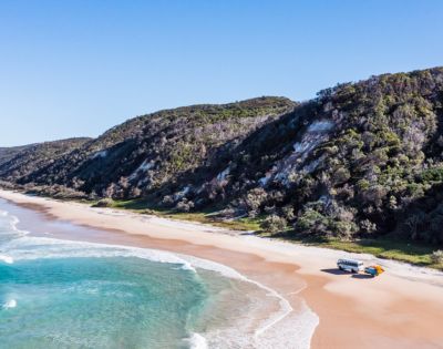 Aerial view of a serene beach with turquoise waves, a camper van and SUV parked on sand, and lush green hills under a clear blue sky.