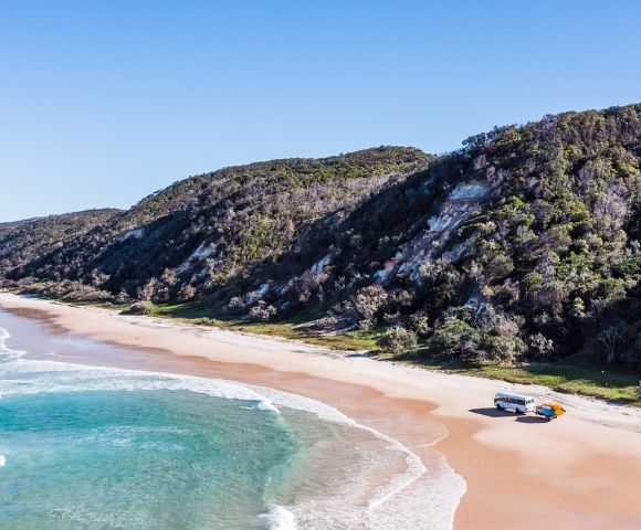 Aerial view of a serene beach with turquoise waves, a camper van and SUV parked on sand, and lush green hills under a clear blue sky.