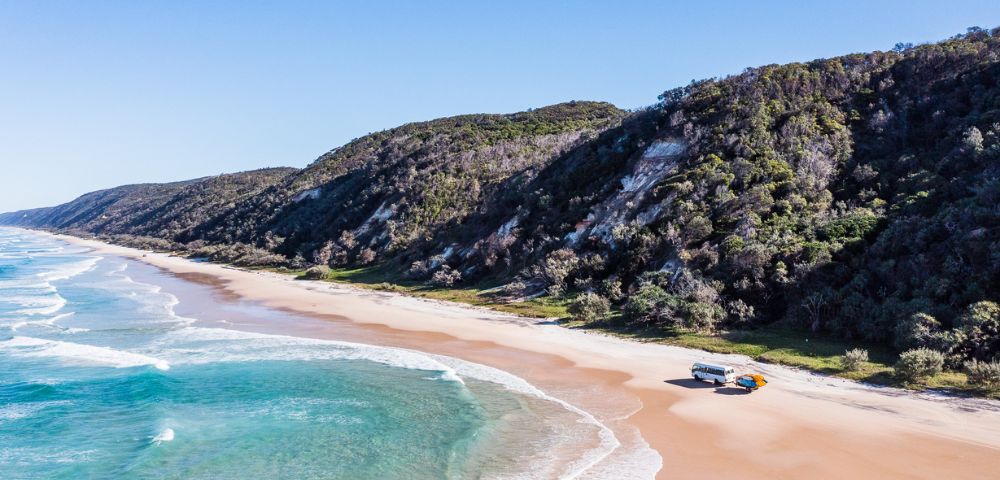 Aerial view of a serene beach with turquoise waves, a camper van and SUV parked on sand, and lush green hills under a clear blue sky.