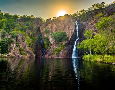 Sunset behind lush cliffs, cascading waterfalls into a serene lake, reflecting golden hues. Verdant trees enhance the tranquil, natural beauty.