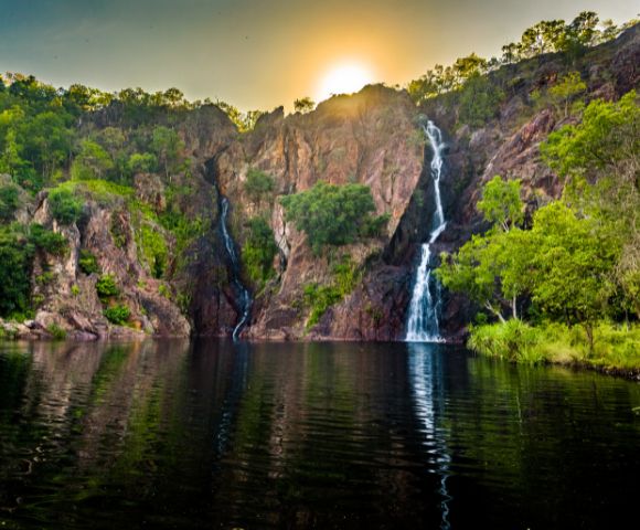 Sunset behind lush cliffs, cascading waterfalls into a serene lake, reflecting golden hues. Verdant trees enhance the tranquil, natural beauty.