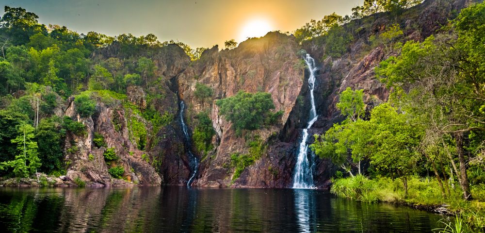 Sunset behind lush cliffs, cascading waterfalls into a serene lake, reflecting golden hues. Verdant trees enhance the tranquil, natural beauty.