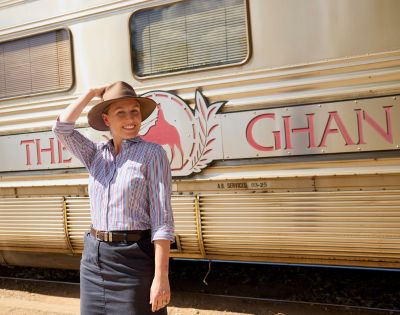 A smiling person in a hat stands in front of a train with 