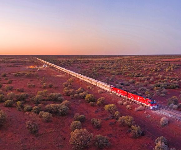 A long train with red locomotives travels through a vast, rugged landscape at sunset, surrounded by sparse vegetation under a softly hued sky.