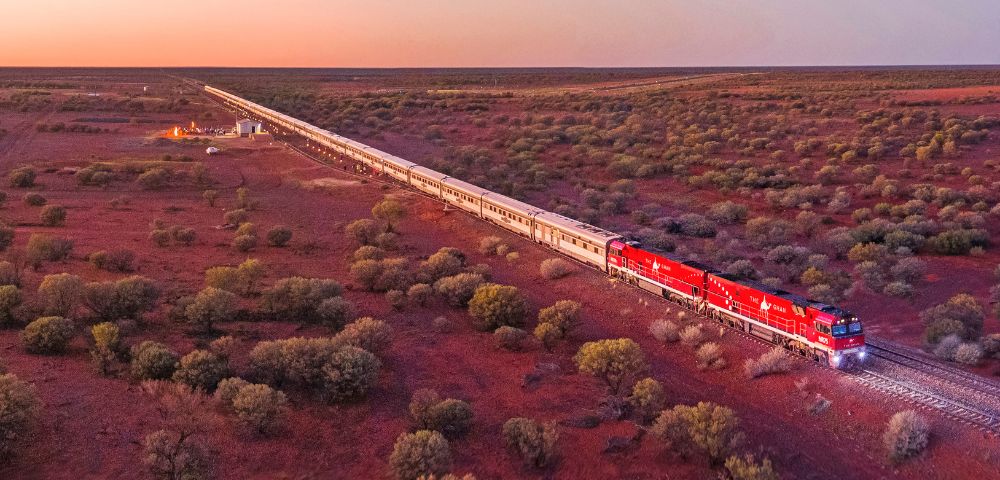 A long train with red locomotives travels through a vast, rugged landscape at sunset, surrounded by sparse vegetation under a softly hued sky.