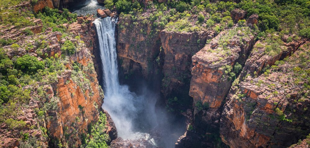 Aerial view of a waterfall cascading into a rocky gorge surrounded by lush green vegetation, conveying a sense of natural beauty and tranquility.