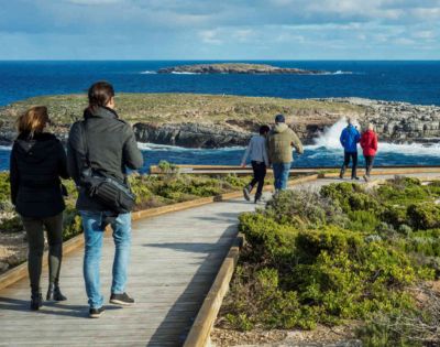 People walk along a wooden boardwalk overlooking a rocky coastline and blue ocean under a partly cloudy sky. The scene is calm and scenic.