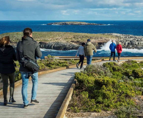 People walk along a wooden boardwalk overlooking a rocky coastline and blue ocean under a partly cloudy sky. The scene is calm and scenic.