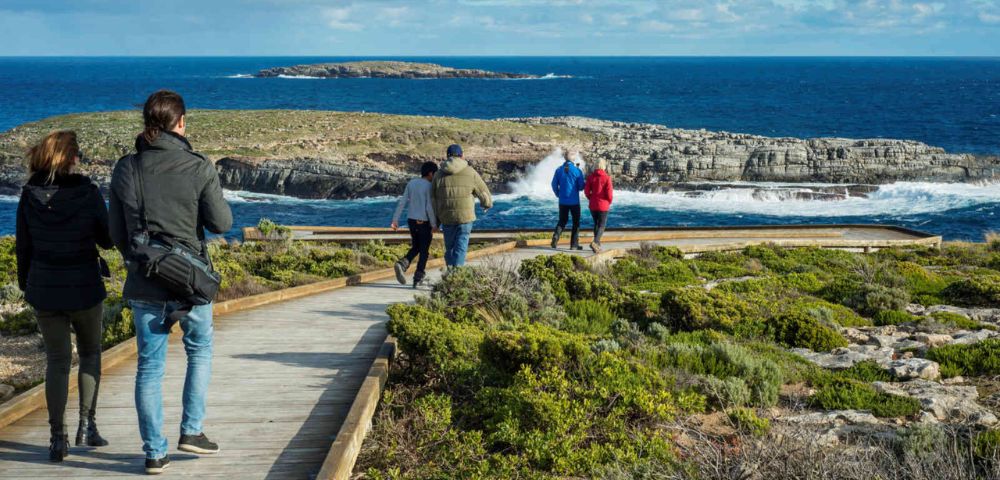 People walk along a wooden boardwalk overlooking a rocky coastline and blue ocean under a partly cloudy sky. The scene is calm and scenic.