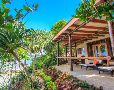 Cozy beachfront bungalow with wooden patio, surrounded by lush greenery. Ocean view, clear blue sky; inviting and tranquil atmosphere.