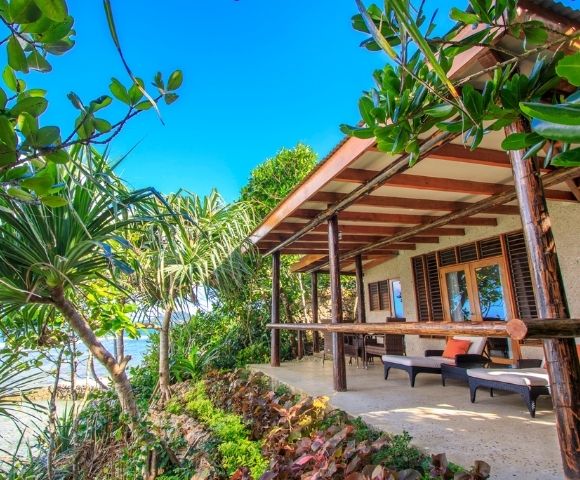 Cozy beachfront bungalow with wooden patio, surrounded by lush greenery. Ocean view, clear blue sky; inviting and tranquil atmosphere.