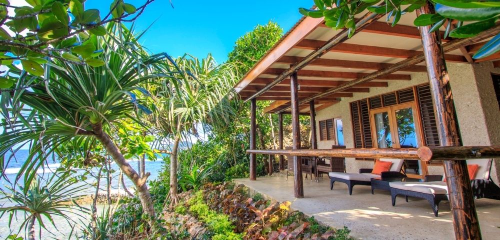 Cozy beachfront bungalow with wooden patio, surrounded by lush greenery. Ocean view, clear blue sky; inviting and tranquil atmosphere.