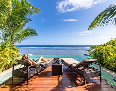 Person relaxing on a lounge chair by an infinity pool, surrounded by tropical plants, gazing at a clear blue ocean under a bright, sunny sky.