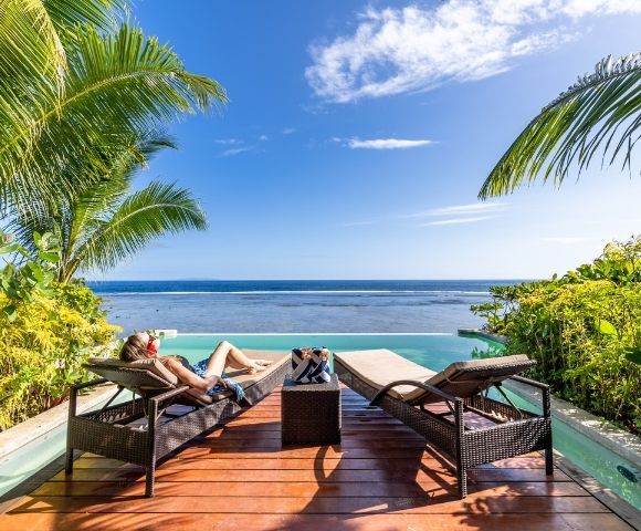 Person relaxing on a lounge chair by an infinity pool, surrounded by tropical plants, gazing at a clear blue ocean under a bright, sunny sky.