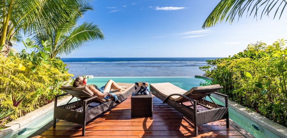 Person relaxing on a lounge chair by an infinity pool, surrounded by tropical plants, gazing at a clear blue ocean under a bright, sunny sky.