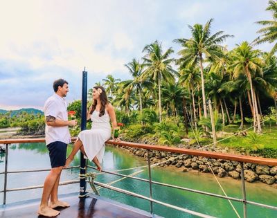 A couple enjoys a tropical moment on a boat near lush palm trees and turquoise water. The scene is serene and romantic, with a warm, joyful atmosphere.