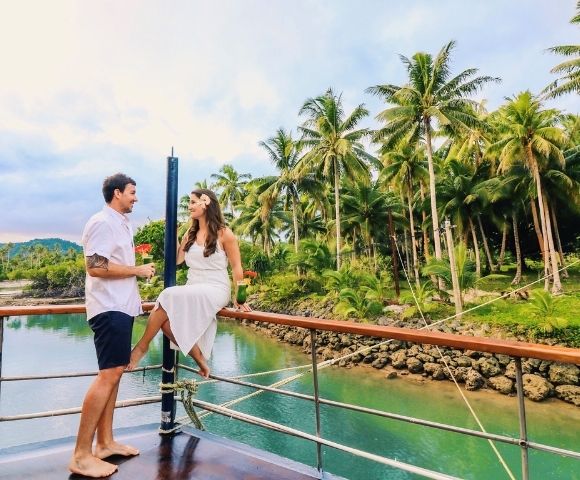 A couple enjoys a tropical moment on a boat near lush palm trees and turquoise water. The scene is serene and romantic, with a warm, joyful atmosphere.