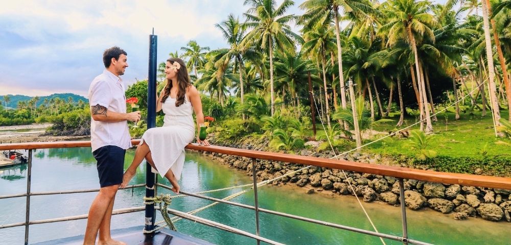 A couple enjoys a tropical moment on a boat near lush palm trees and turquoise water. The scene is serene and romantic, with a warm, joyful atmosphere.