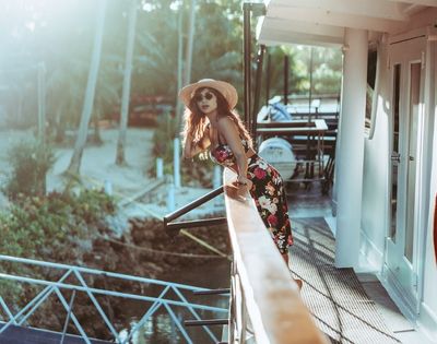 A woman in a floral dress and sun hat leans over a boat railing, gazing at the sunny beach lined with palm trees, conveying a relaxed, summery vibe.