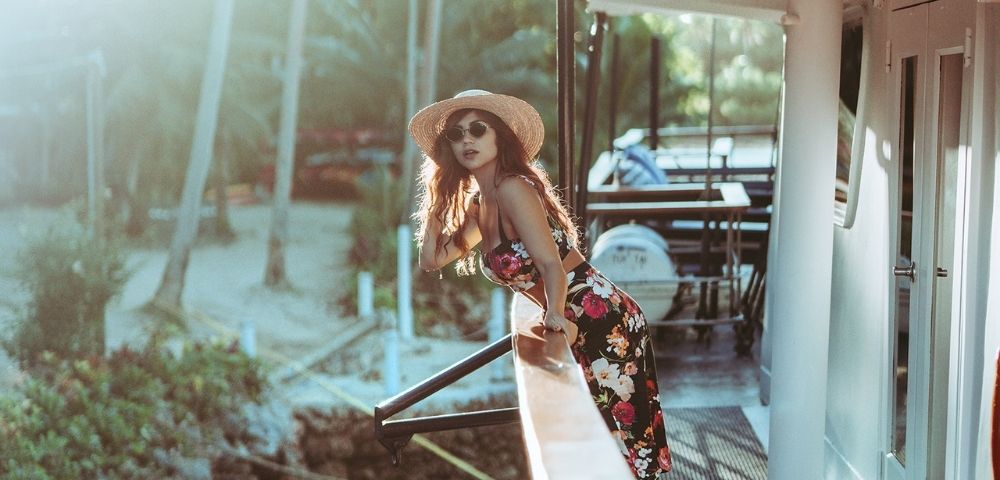 A woman in a floral dress and sun hat leans over a boat railing, gazing at the sunny beach lined with palm trees, conveying a relaxed, summery vibe.
