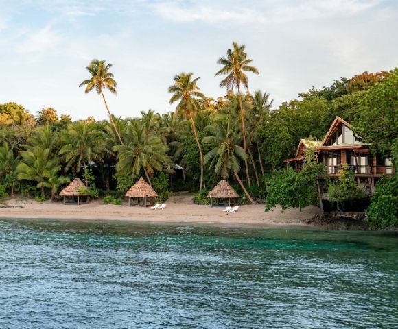 A tropical beach scene with palm trees, three thatched umbrellas on a sandy shore, and a wooden beach house surrounded by lush greenery by the calm sea.