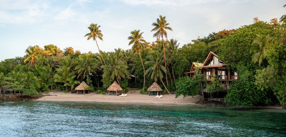A tropical beach scene with palm trees, three thatched umbrellas on a sandy shore, and a wooden beach house surrounded by lush greenery by the calm sea.