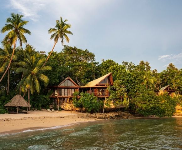 Tropical beach scene with lush greenery and palm trees surrounding charming wooden huts. A tranquil ocean meets the sandy shore under a bright sky.