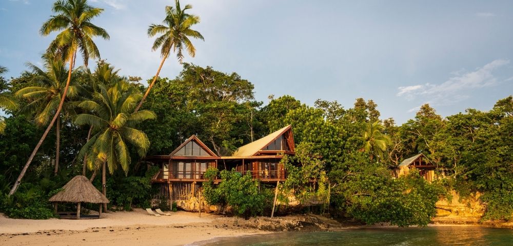 Tropical beach scene with lush greenery and palm trees surrounding charming wooden huts. A tranquil ocean meets the sandy shore under a bright sky.