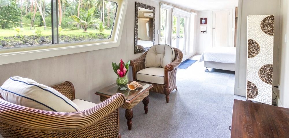 A cozy living room with wicker chairs and a small table with flowers, facing a large window showing lush greenery. A bedroom is visible in the background.