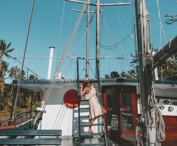 Woman in a beige dress and wide-brimmed hat stands on a boat ladder, posing confidently. Palm trees in the background add a tropical feel. Sunny day.