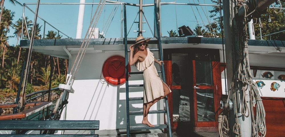 Woman in a beige dress and wide-brimmed hat stands on a boat ladder, posing confidently. Palm trees in the background add a tropical feel. Sunny day.