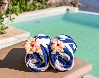Two rolled blue-and-white striped towels with hibiscus flowers rest on a sunbed by a serene pool, surrounded by lush greenery and a calm atmosphere.