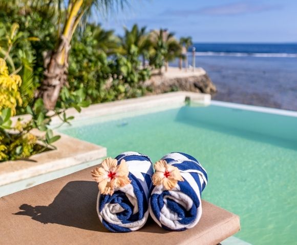 Two rolled blue-and-white striped towels with hibiscus flowers rest on a sunbed by a serene pool, surrounded by lush greenery and a calm atmosphere.