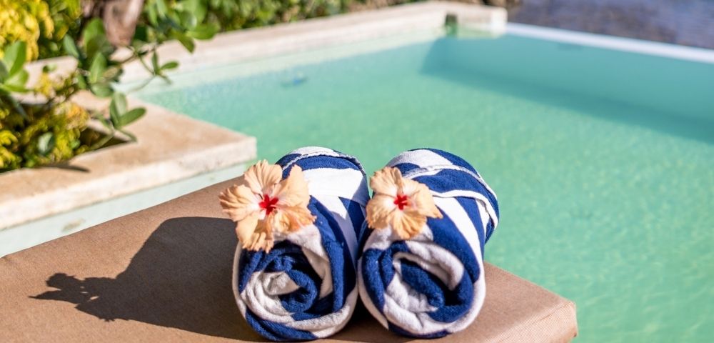 Two rolled blue-and-white striped towels with hibiscus flowers rest on a sunbed by a serene pool, surrounded by lush greenery and a calm atmosphere.