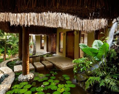 Thatched-roof tropical villa surrounded by lush greenery. A serene pond with lily pads and stone pathways create a peaceful, natural ambiance.