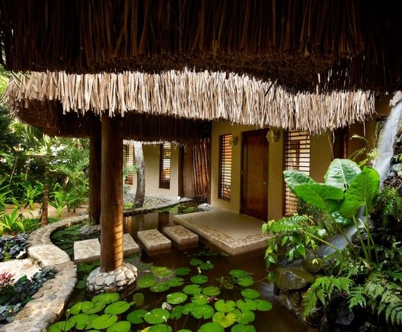 Thatched-roof tropical villa surrounded by lush greenery. A serene pond with lily pads and stone pathways create a peaceful, natural ambiance.