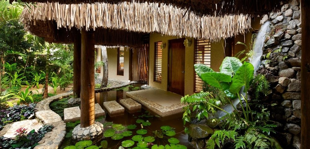 Thatched-roof tropical villa surrounded by lush greenery. A serene pond with lily pads and stone pathways create a peaceful, natural ambiance.