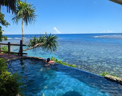 A person relaxes in an infinity pool overlooking a calm, clear ocean. Tropical plants surround the pool, creating a serene and peaceful atmosphere.