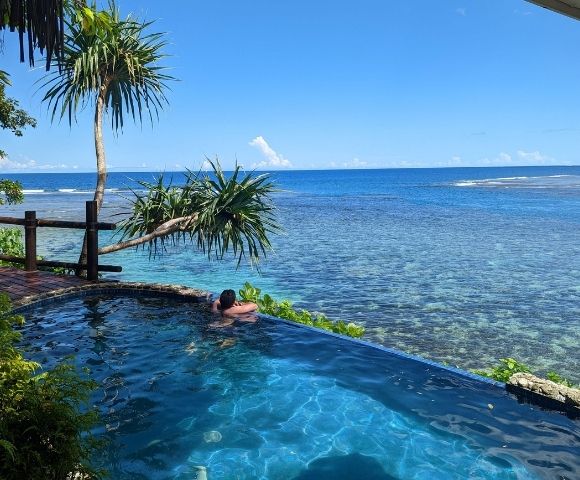 A person relaxes in an infinity pool overlooking a calm, clear ocean. Tropical plants surround the pool, creating a serene and peaceful atmosphere.