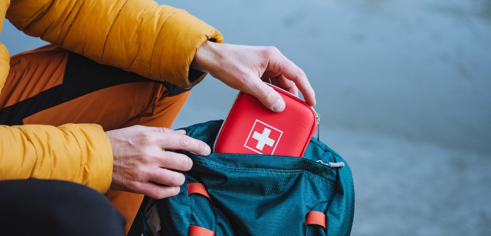 Hands placing a red first aid kit with a white cross into the front pocket of a teal backpack, with a yellow jacket sleeve visible.